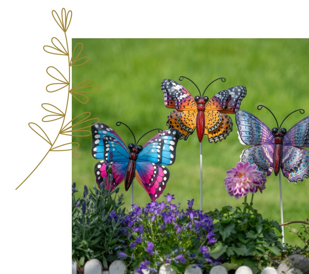 formano.2025.bildmaterial.garten.571786 Drei bunte Dekoschmetterlinge in leuchtenden Farben (Blau, Rot-Orange und Lila) sind auf Metallstäben in einem blühenden Garten befestigt. Im Vordergrund wachsen violette Glockenblumen und Dahlien, während ein kleiner weißer Lattenzaun die Szene umrahmt. Im Hintergrund erstreckt sich eine grüne Wiese. Ein stilisiertes Pflanzenmotiv auf der linken Seite des Bildes ergänzt das Gesamtbild und verleiht ihm eine kreative, dekorative Note. Die Szene wirkt lebendig, fröhlich und sommerlich.